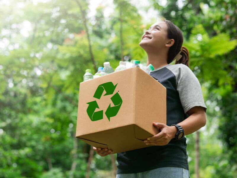 Pessoa carregando caixa de reciclagem com garrafas plásticas em ambiente verde